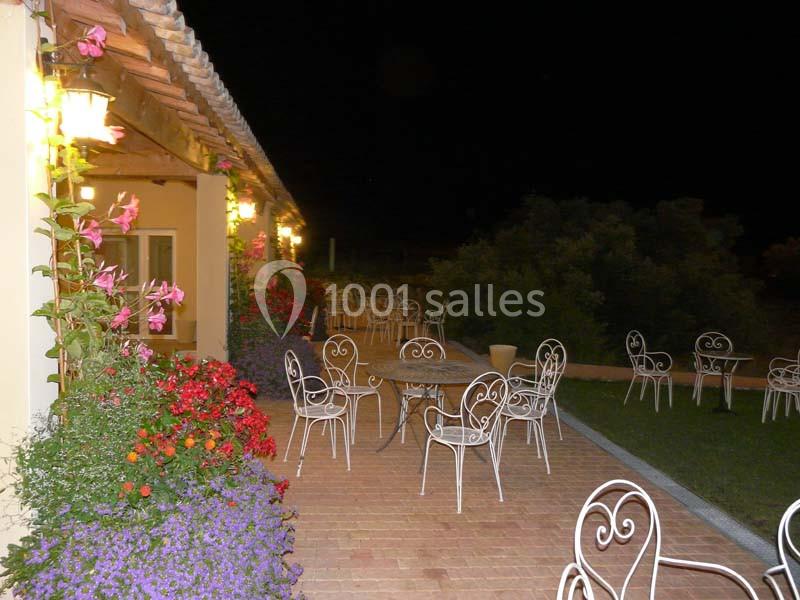 Terrasse extérieure éclairée la nuit, avec des tables et chaises en fer forgé entourées de fleurs colorées.