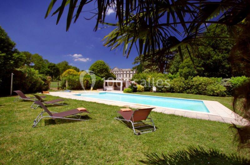 Piscine extérieure entourée de chaises longues sur une pelouse, avec un bâtiment en arrière-plan et des arbres autour.