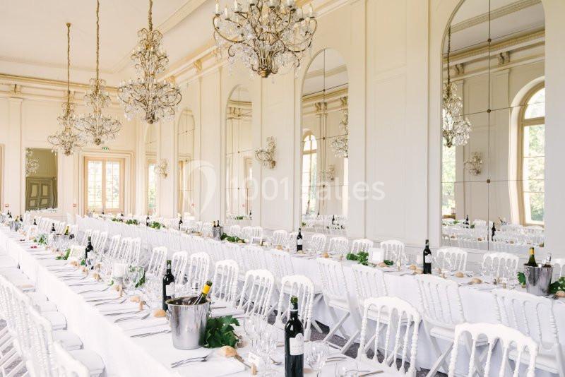 Salle de réception élégante avec tables longues dressées, chaises blanches et lustres suspendus.