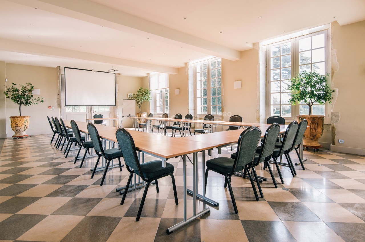 Salle lumineuse aménagée en configuration de réunion avec tables en U, chaises noires et grand écran de projection.
