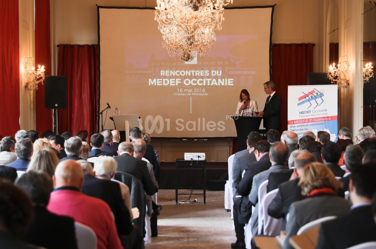 Conférence avec des intervenants sur scène devant un public, dans une salle ornée de lustres et de rideaux rouges.