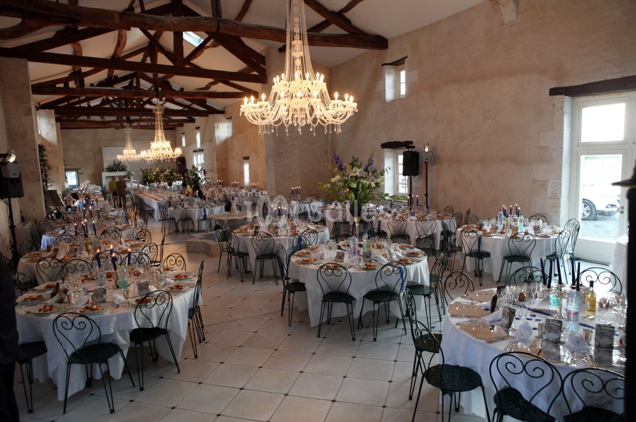 Salle de réception lumineuse avec tables rondes dressées, chaises en fer forgé et lustres suspendus.