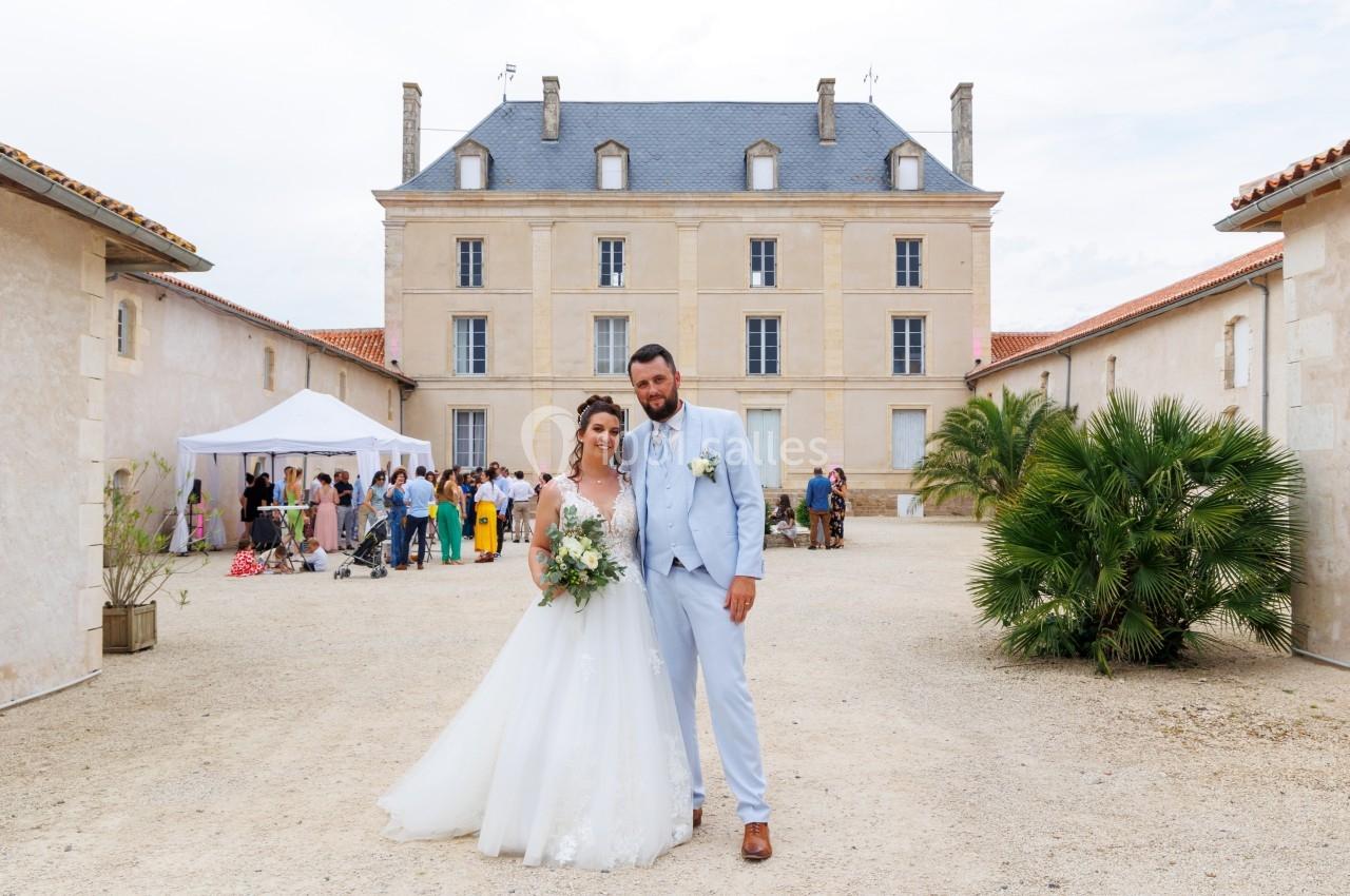 Un couple en tenue de mariage pose devant un bâtiment historique entouré d'invités et de végétation.