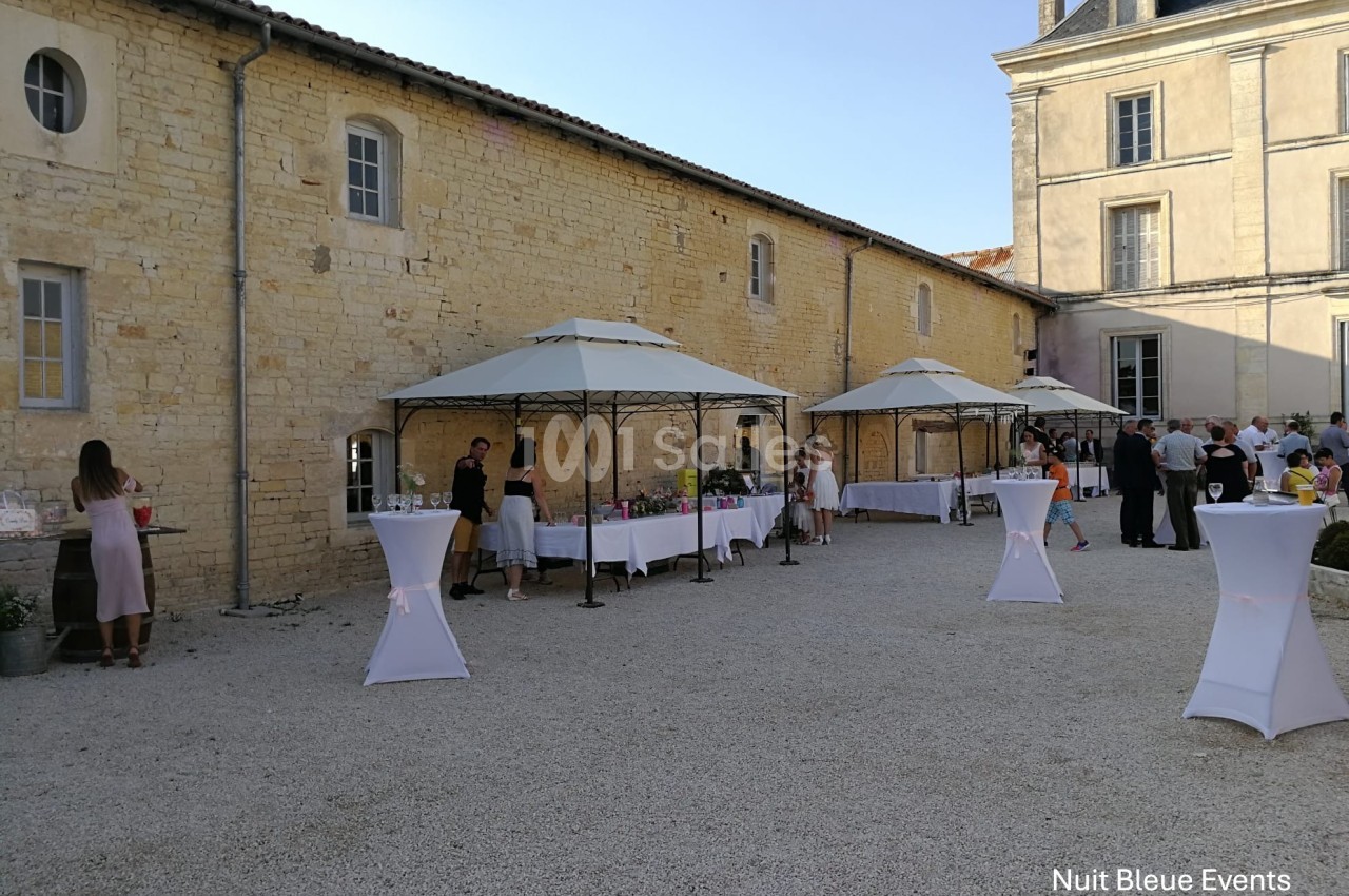 Cour extérieure avec des tables sous des tonnelles, des invités debout et un bâtiment en pierre en arrière-plan.