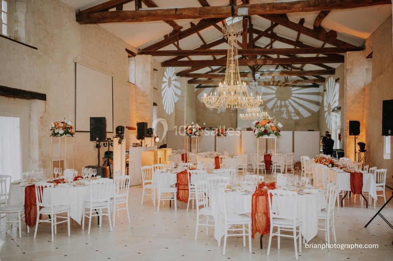 Salle de réception élégante avec tables décorées, chaises blanches, lustres et poutres apparentes en bois.