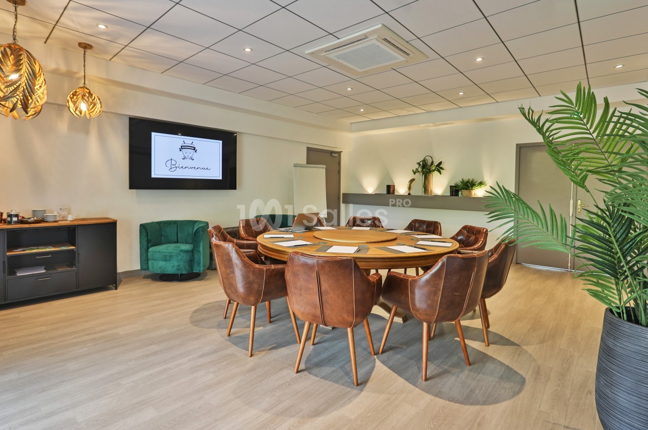 Salle de réunion lumineuse avec table ronde, chaises en cuir, écran mural et plantes décoratives.