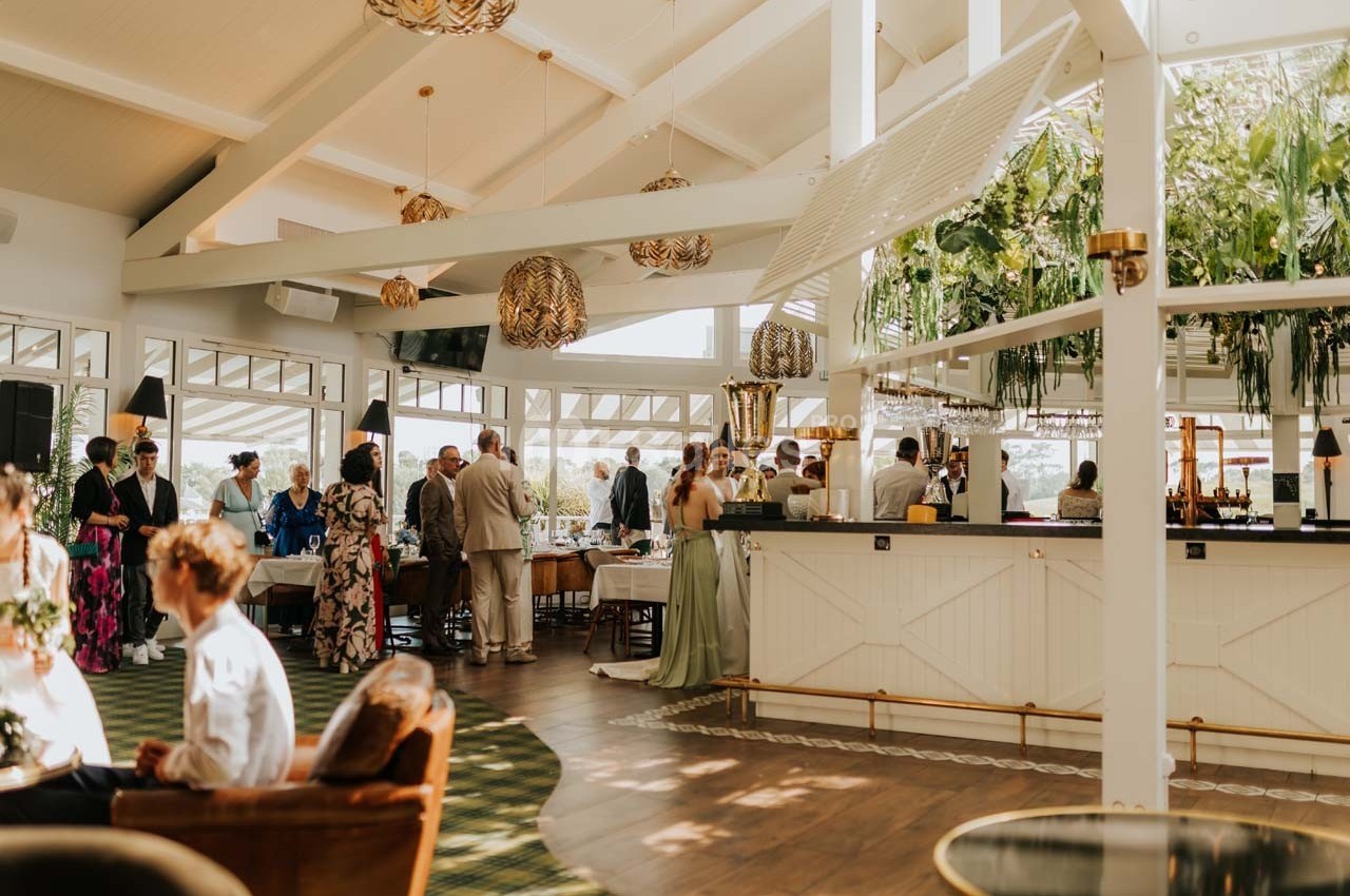 Salle lumineuse avec des invités debout et assis, décorée de plantes suspendues et d'un bar central.