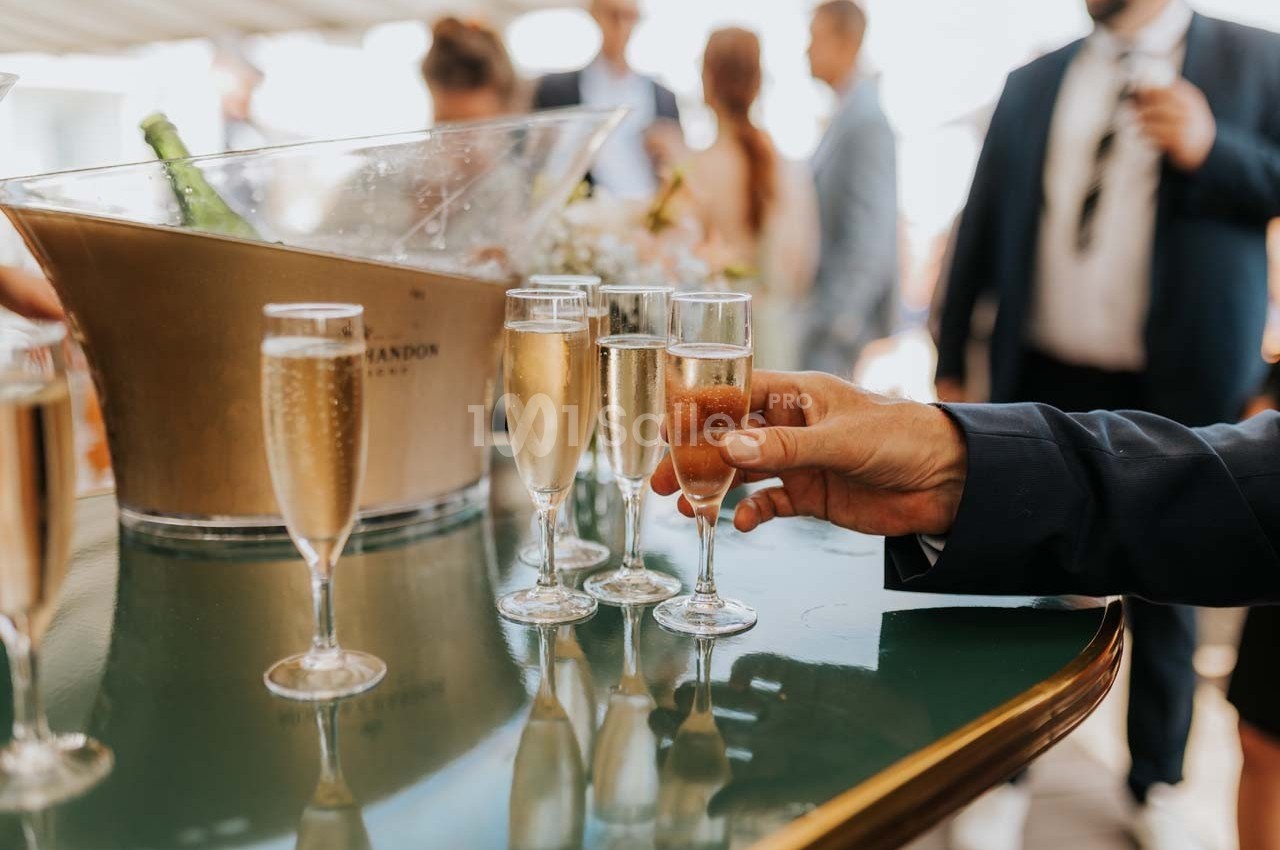 Un homme en costume prend une flûte de champagne sur une table avec d'autres verres et un seau à champagne en arrière-plan.