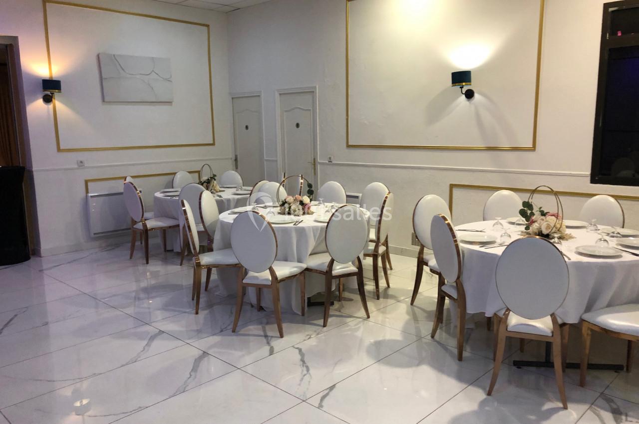 Salle de réception lumineuse avec tables rondes dressées, chaises blanches et décorations florales.