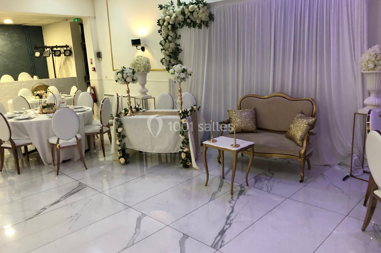 Salle de réception décorée avec des tables rondes, un espace d'honneur avec arche florale et un canapé doré.