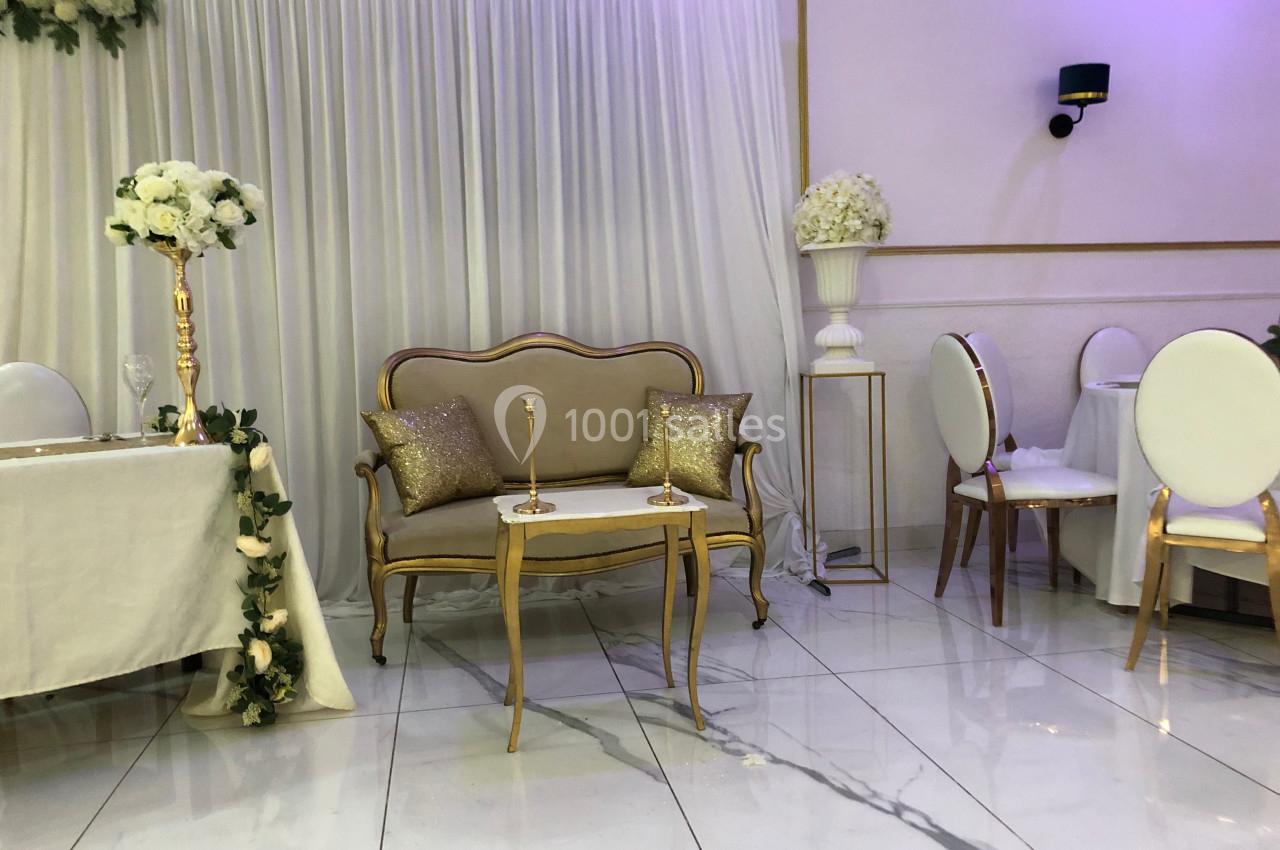 Salle décorée pour un événement avec canapé doré, table assortie, chaises blanches et arrangements floraux blancs.