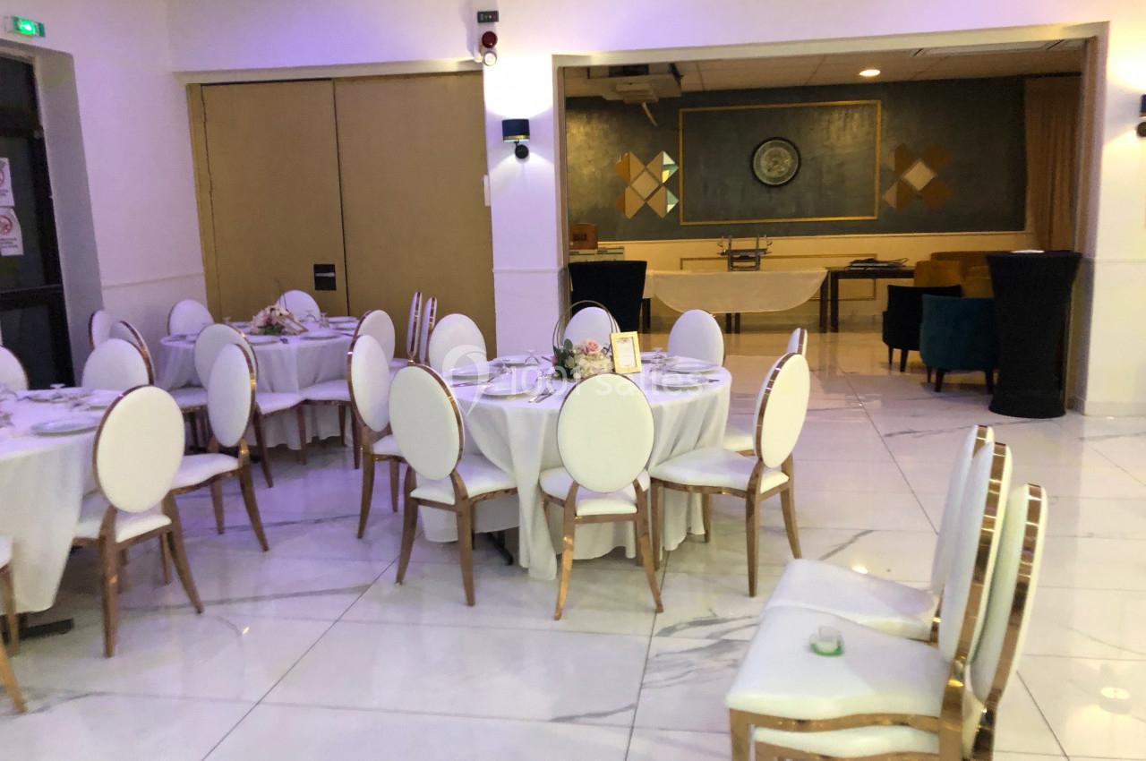 Salle de réception lumineuse avec tables rondes dressées, chaises blanches et décorations sobres.