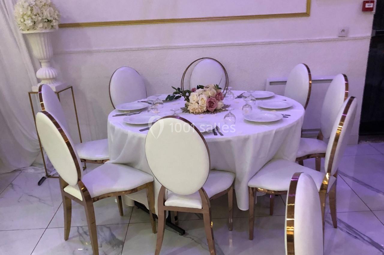 Table ronde dressée avec une nappe blanche, entourée de chaises blanches, décorée d'un bouquet de fleurs au centre.