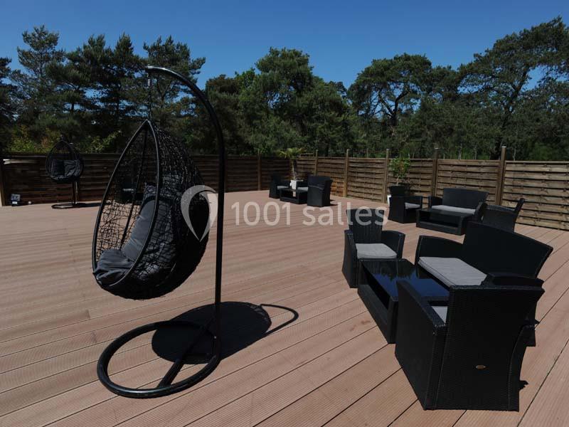 Terrasse en bois avec fauteuils en résine tressée et fauteuil suspendu, entourée de panneaux en bois et arbres en arrière…