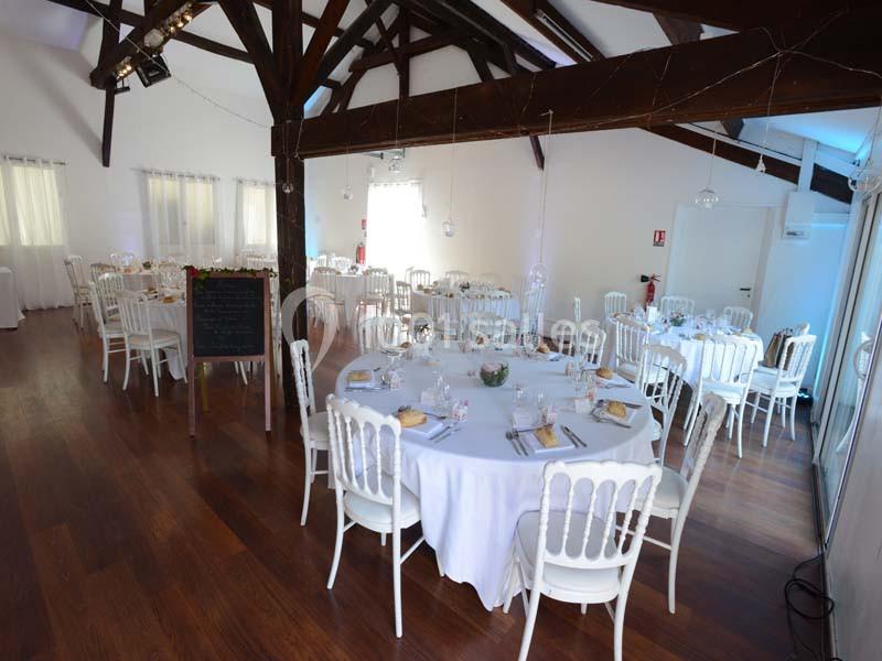 Salle lumineuse avec tables rondes dressées, chaises blanches, parquet en bois et poutres apparentes.