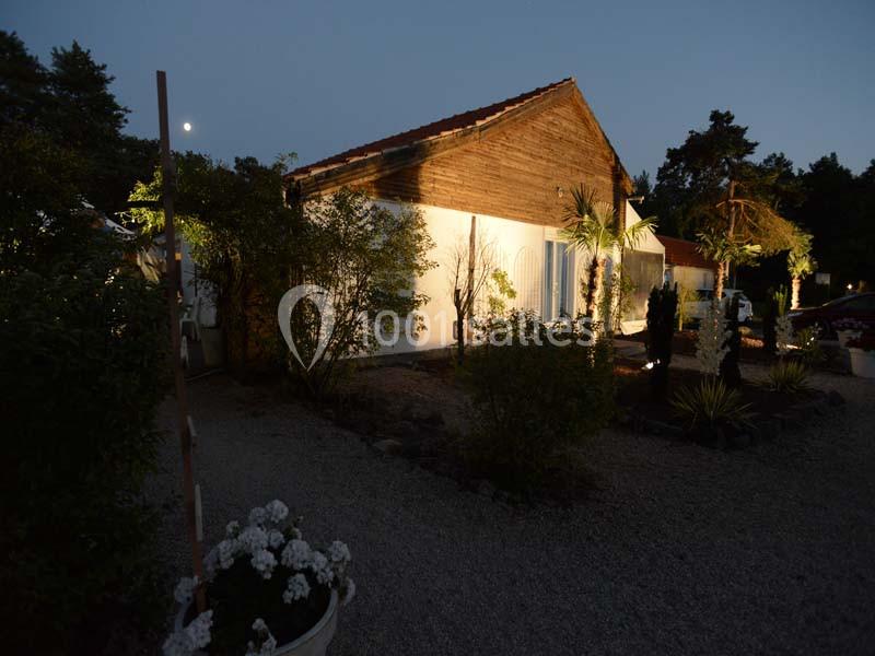 Maison en bois et crépi blanc éclairée la nuit, entourée de végétation et d'un jardin avec graviers.