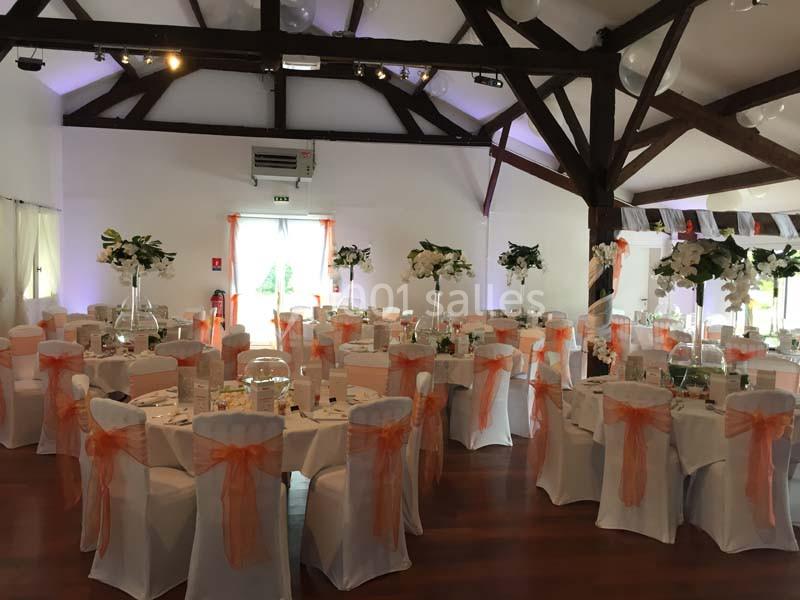Salle de réception décorée pour un événement, avec des chaises blanches ornées de nœuds orange et des tables dressées.