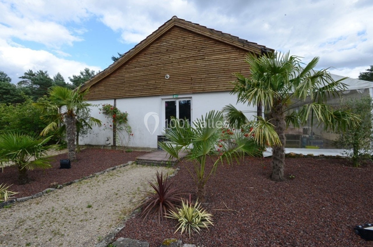 Façade d'une maison avec un toit en bois, entourée de palmiers et d'un jardin aménagé avec du gravier rouge.