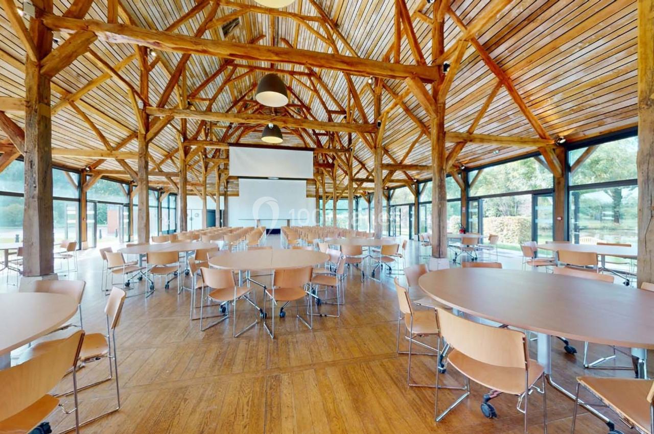 Salle lumineuse avec charpente en bois, tables rondes et chaises disposées face à un écran de projection.