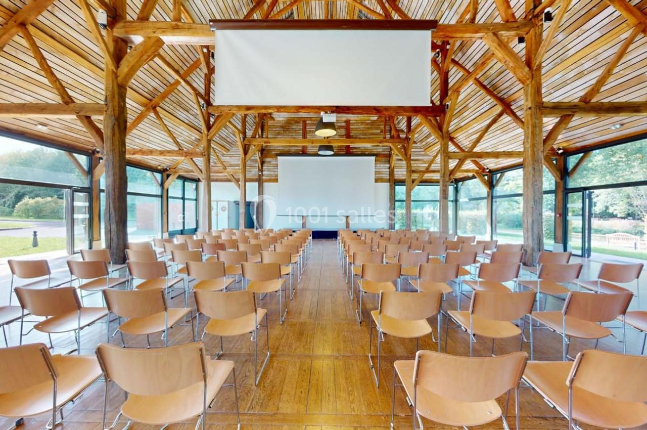 Salle de conférence lumineuse avec des rangées de chaises en bois, écrans suspendus et grandes baies vitrées donnant sur l…