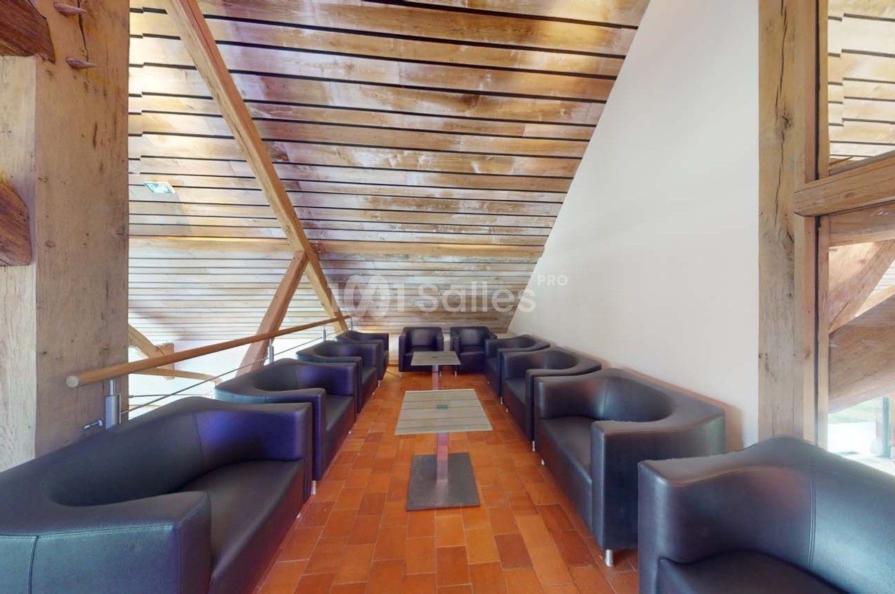 Espace lounge avec fauteuils noirs disposés autour de petites tables sur un sol en carreaux rouges, sous un plafond en bois.