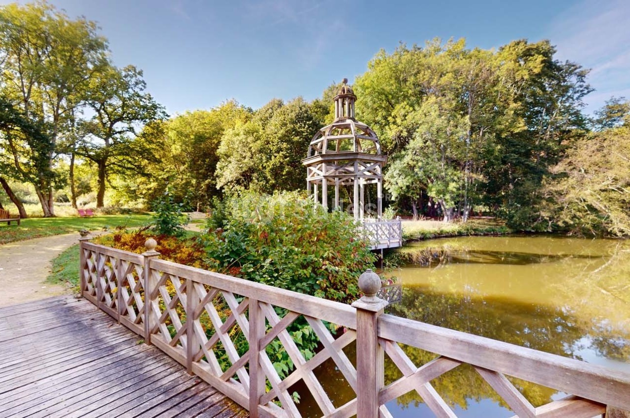 Pont en bois menant à un kiosque au bord d'un étang entouré d'arbres sous un ciel dégagé.