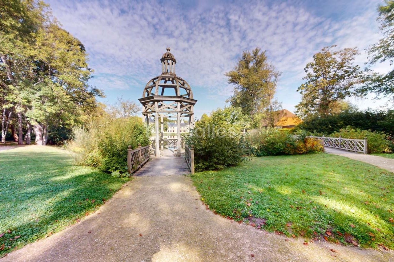 Kiosque en bois au centre d'un jardin verdoyant, entouré de chemins et de végétation sous un ciel partiellement nuageux.