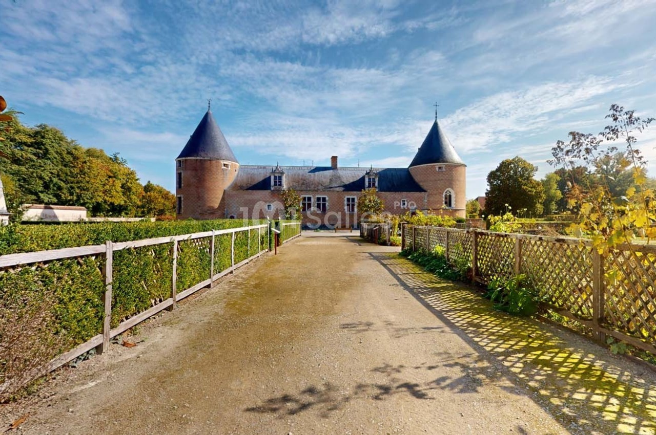 Allée bordée de haies menant à un château en briques rouges avec deux tours, sous un ciel dégagé.