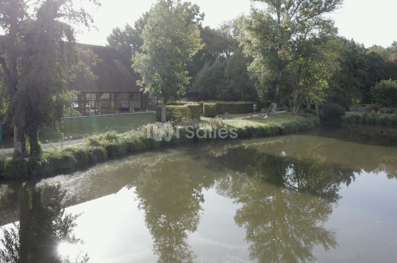 Plan d'eau calme bordé d'arbres et d'une maison à toit en chaume, reflétant le paysage environnant.