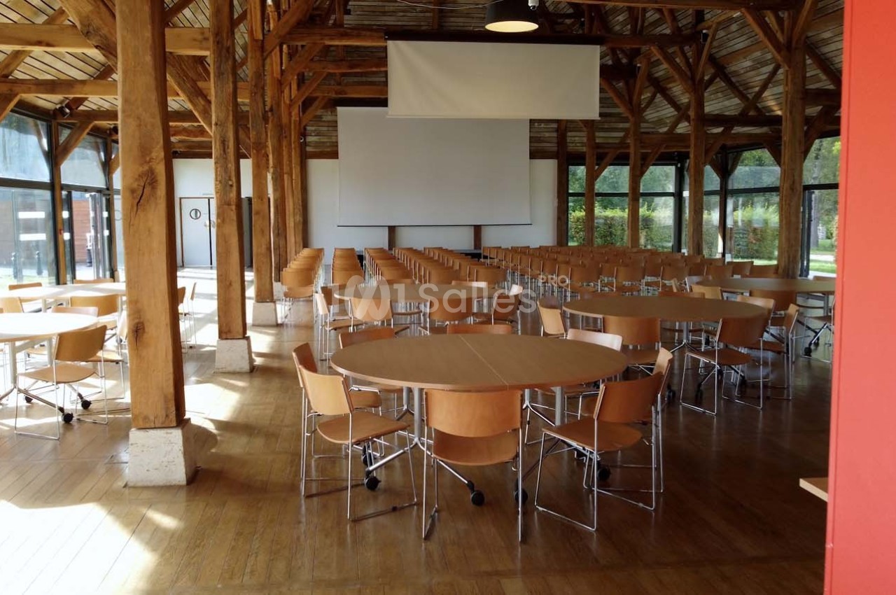 Salle de réunion lumineuse avec des tables rondes en bois, des chaises et un écran de projection au fond.