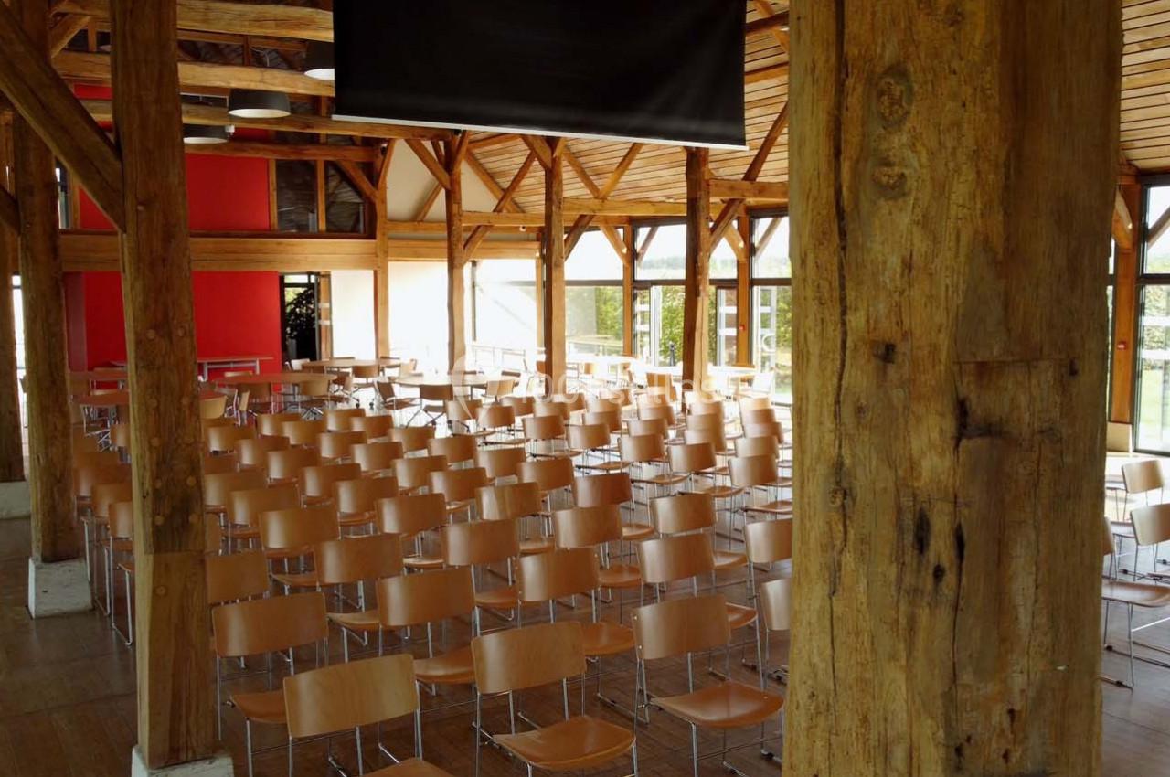 Salle de conférence lumineuse avec des rangées de chaises en bois clair, poutres apparentes et grandes fenêtres.