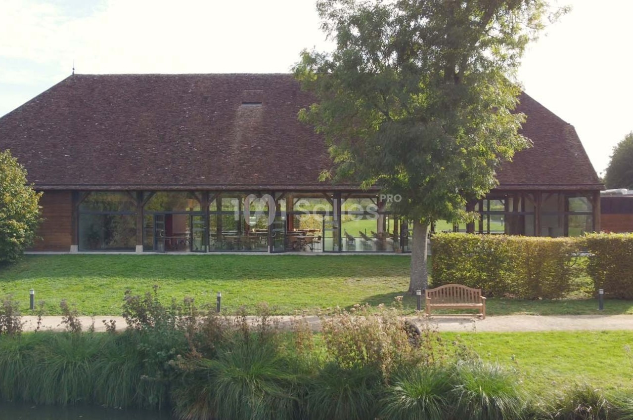 Bâtiment en bois avec de grandes baies vitrées, entouré de verdure et d'un chemin longeant un étang.
