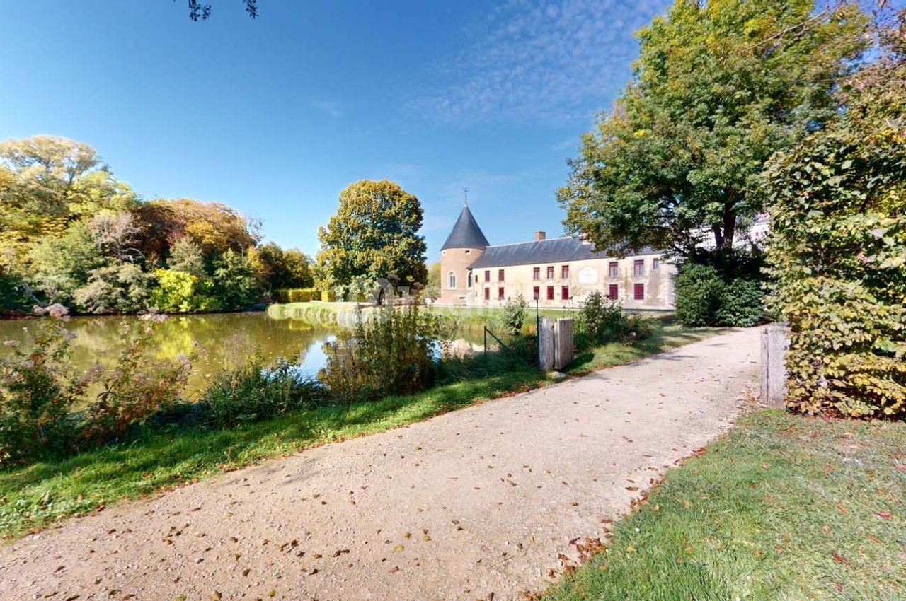 Chemin bordé de verdure menant à un bâtiment en pierre avec une tour, près d'un étang entouré d'arbres.