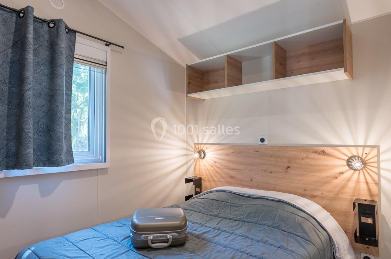 Chambre lumineuse avec lit double, tête de lit en bois, valise posée, étagères ouvertes et fenêtre avec rideaux bleus.