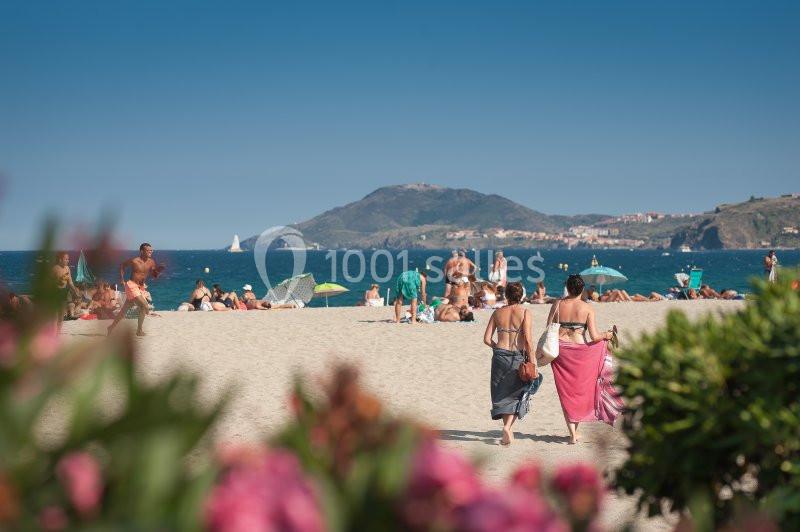 Deux femmes marchent sur une plage ensoleillée, entourées de vacanciers, avec une colline en arrière-plan.