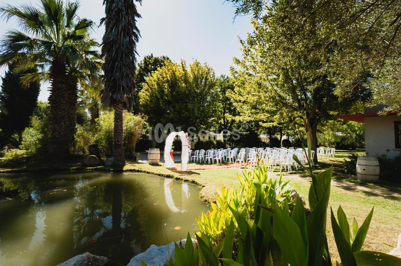 Un jardin avec un étang, des chaises blanches disposées pour une cérémonie et une arche décorative sous les arbres.