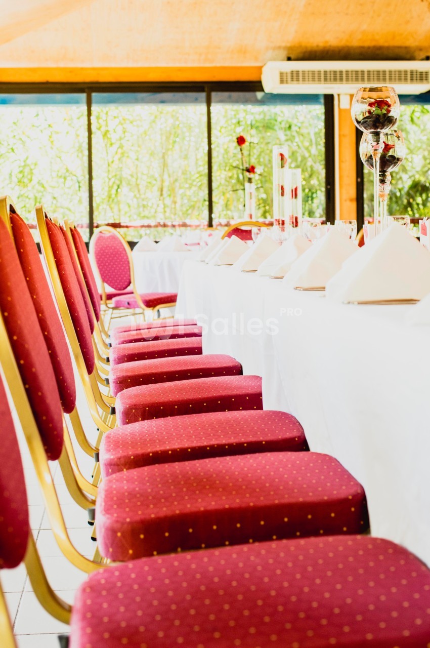 Chaises rouges alignées le long d'une table dressée avec nappes blanches et décorations dans une salle lumineuse.