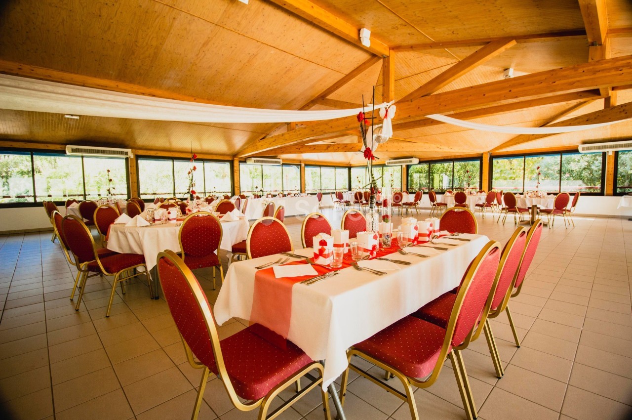 Salle de réception lumineuse avec tables dressées, nappes blanches, chaises rouges et décorations festives.