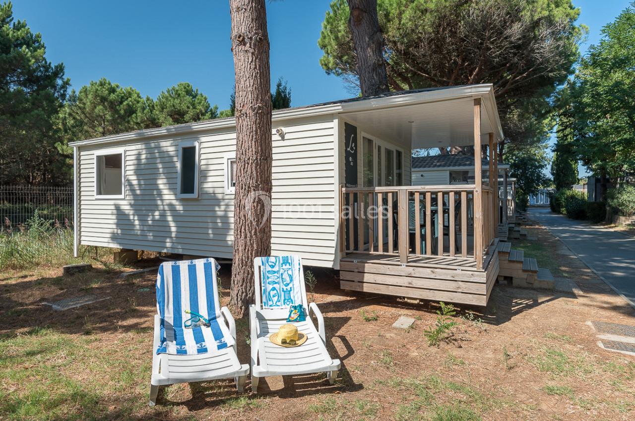 Mobil-home avec terrasse en bois entouré d'arbres, deux chaises longues au premier plan sur un sol en terre.