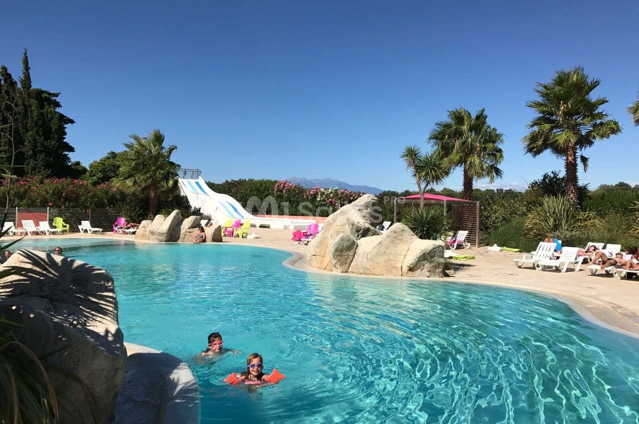 Piscine extérieure avec toboggans, palmiers et transats, deux personnes nageant sous un ciel dégagé.