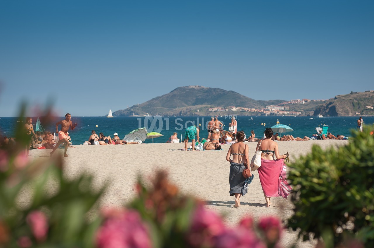 Deux femmes marchant sur une plage ensoleillée, avec des baigneurs et une colline en arrière-plan.