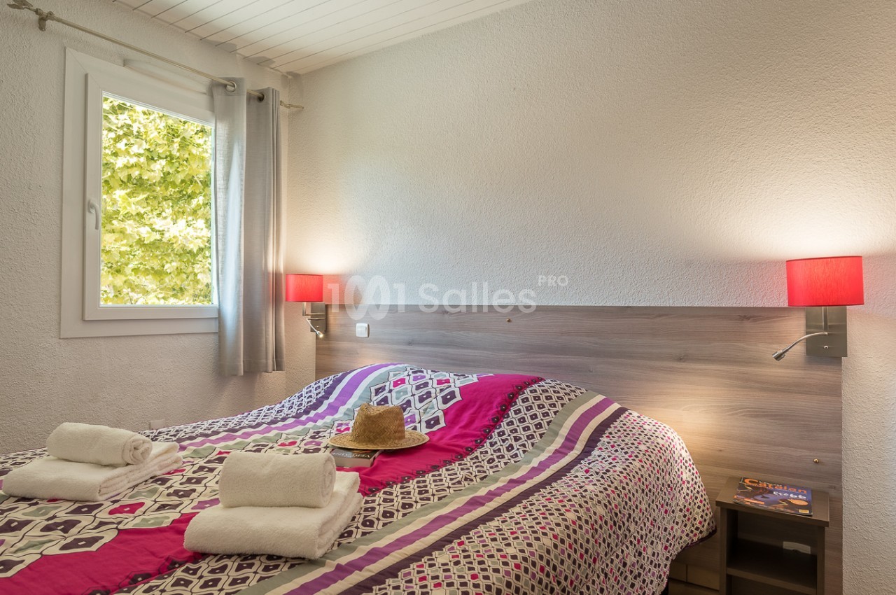 Chambre lumineuse avec lit double, dessus-de-lit coloré, serviettes pliées et fenêtre donnant sur des arbres.
