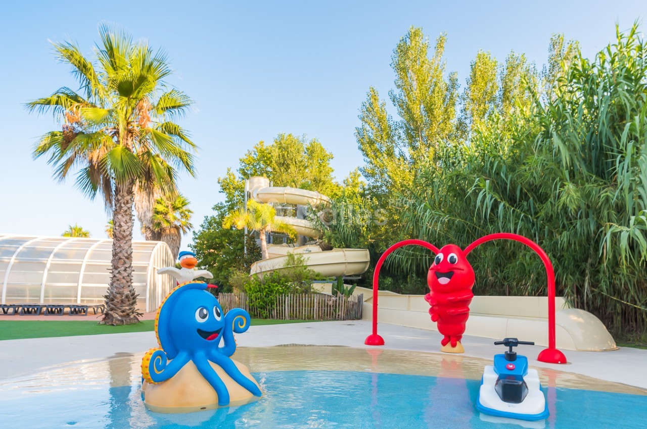 Piscine pour enfants avec jeux aquatiques colorés, toboggan en arrière-plan et végétation environnante.