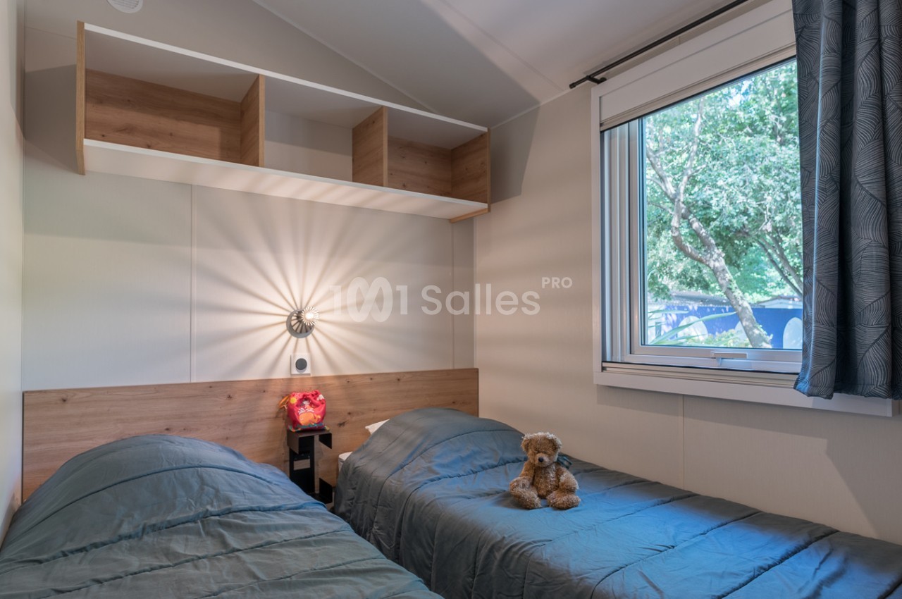 Chambre avec deux lits simples, une lampe murale allumée, une fenêtre donnant sur des arbres et une peluche sur un lit.