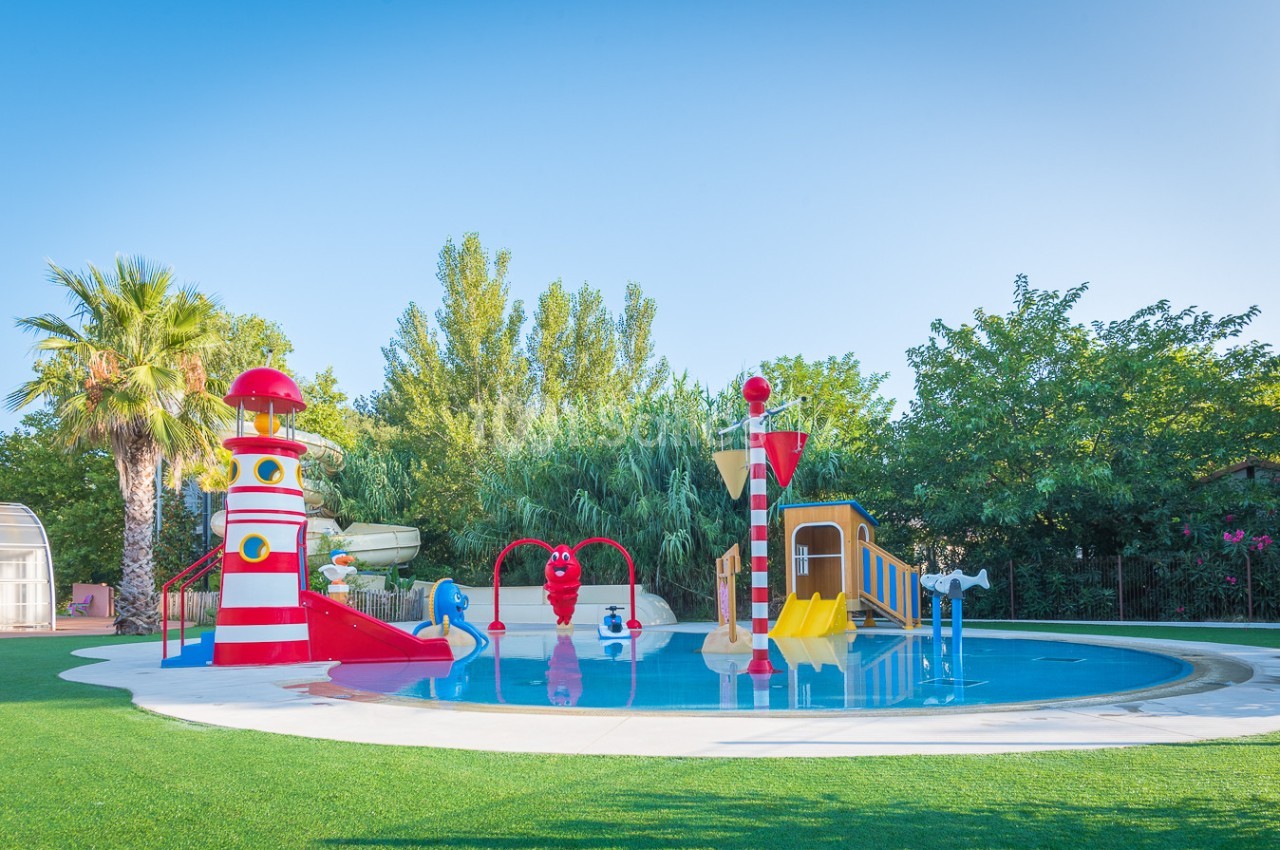 Aire de jeux aquatique avec toboggans colorés, jets d'eau et décorations en forme de phare, entourée de verdure.