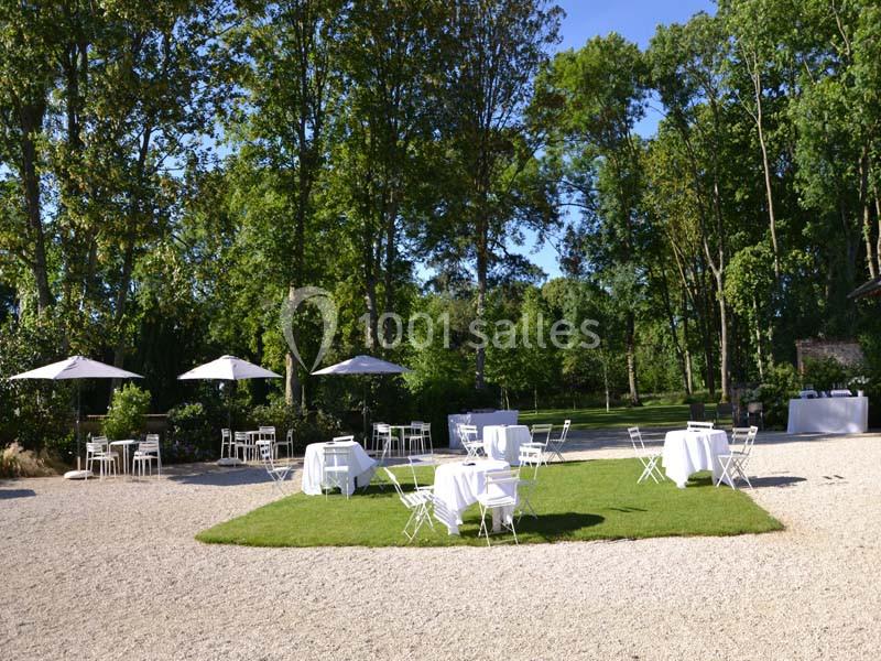 Espace extérieur aménagé avec tables et chaises blanches sur une pelouse entourée d'arbres, sous un ciel dégagé.
