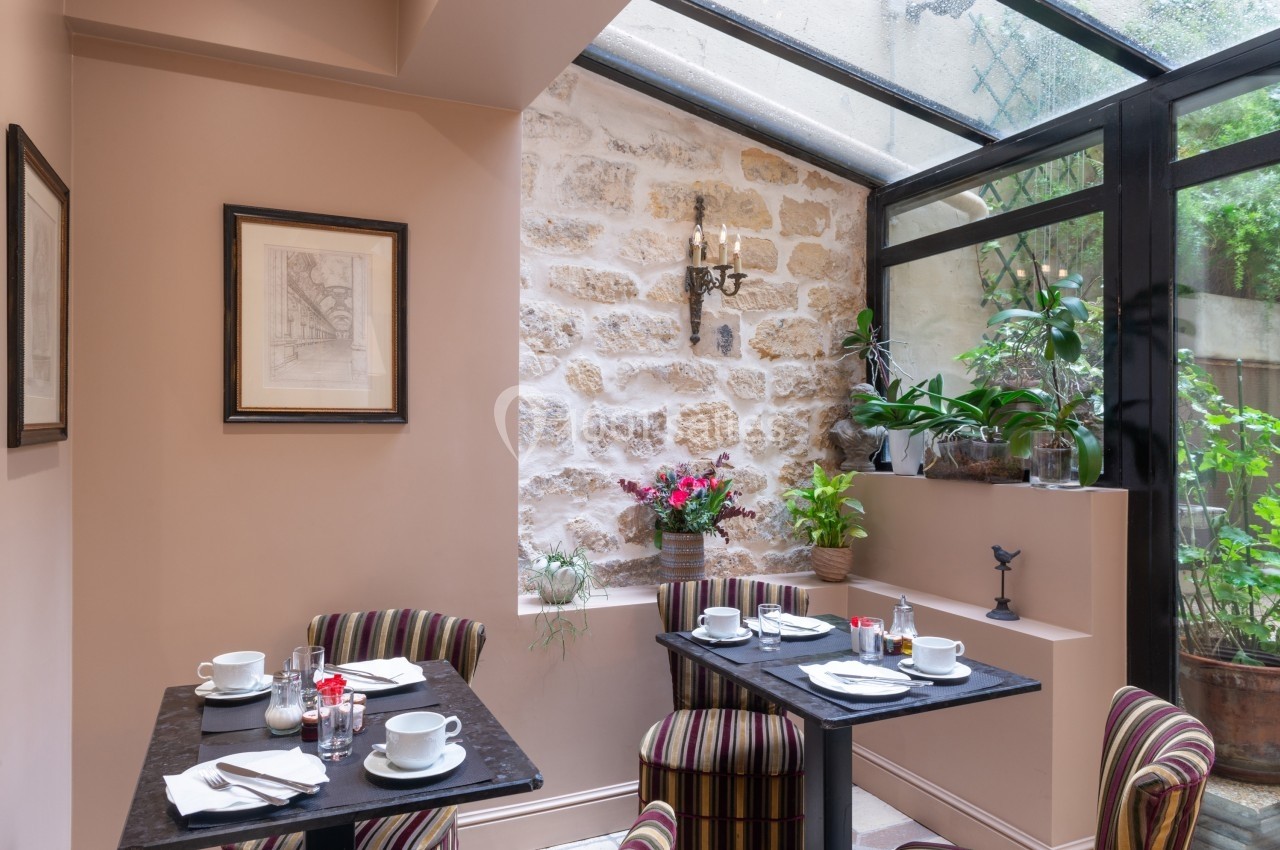 Salle de restaurant lumineuse avec murs en pierre, verrière, plantes et tables dressées pour le petit-déjeuner.
