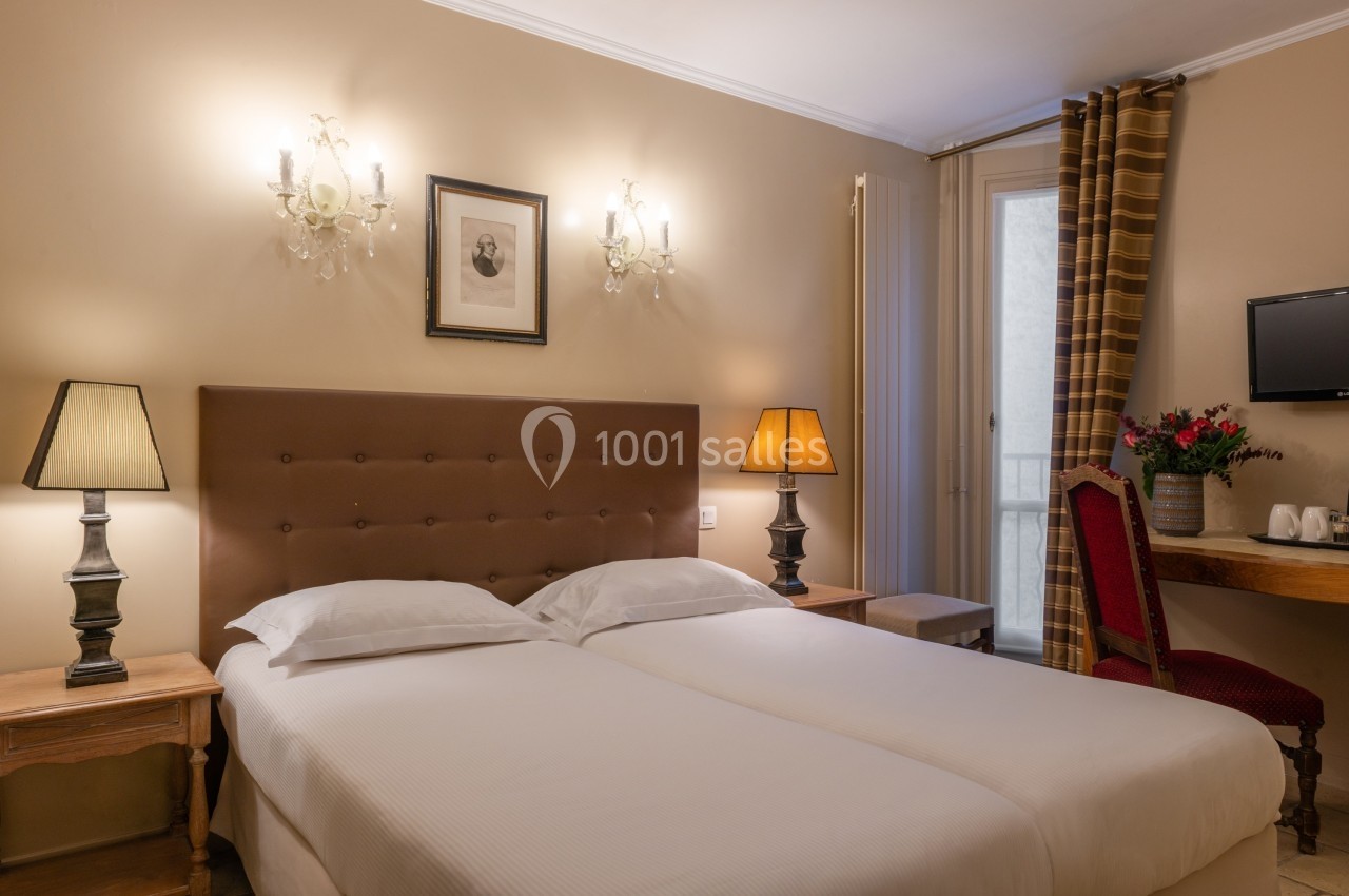 Chambre avec deux lits simples, éclairée par des lampes de chevet, avec un bureau, une chaise et une télévision murale.