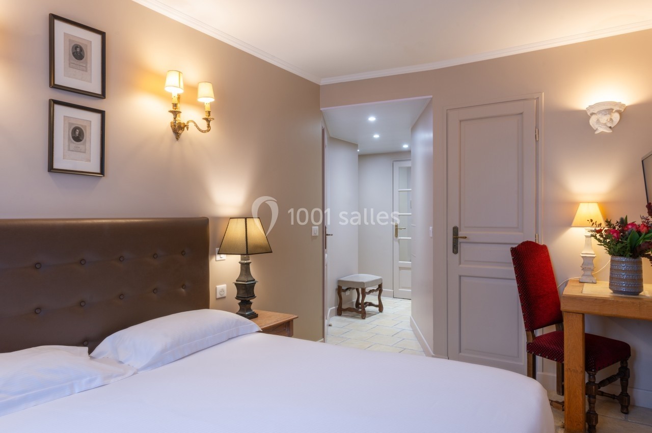 Chambre avec lit double, bureau en bois, chaise rouge, éclairage doux et vue sur un couloir carrelé.