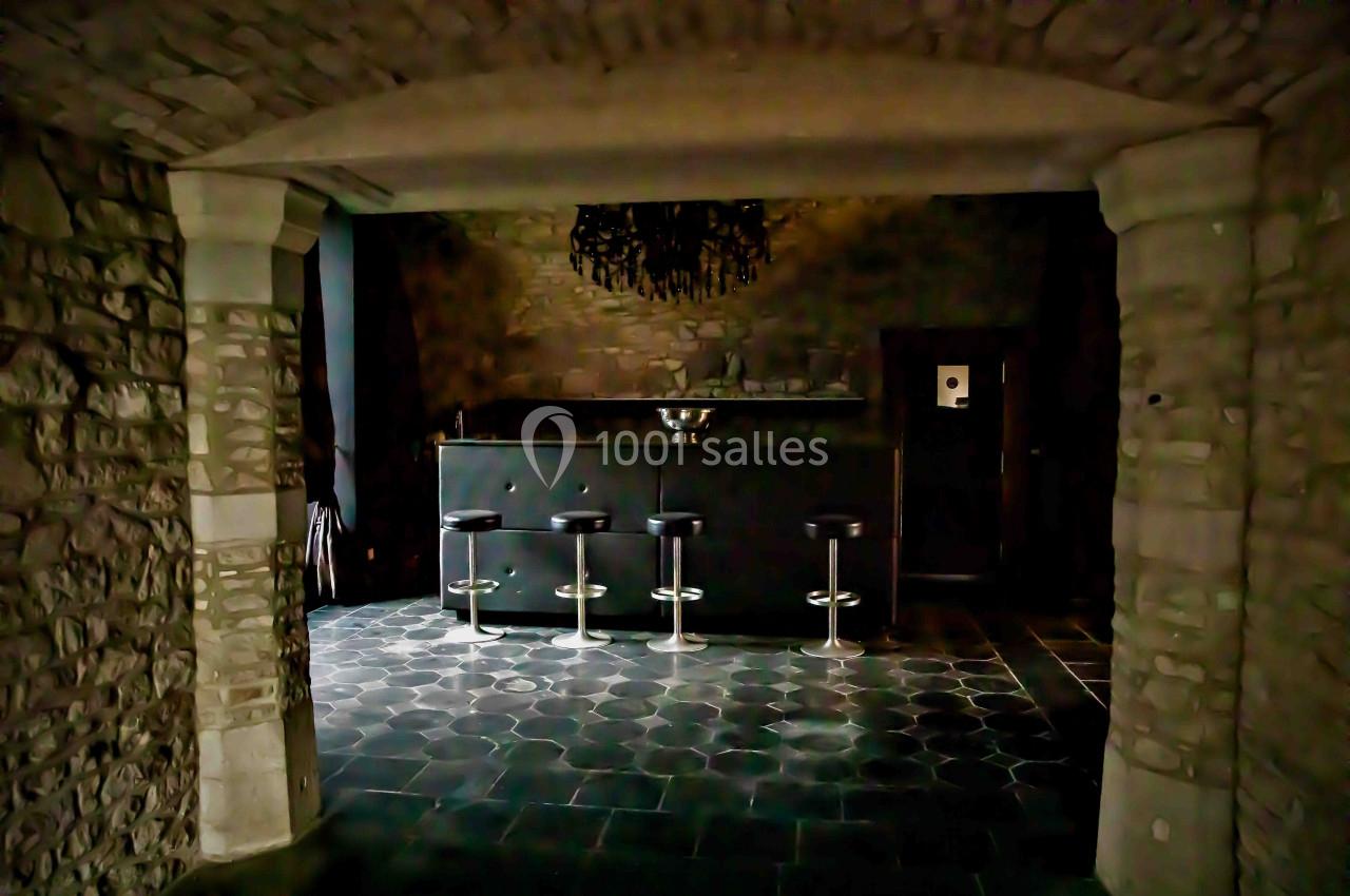 Bar en pierre avec comptoir sombre, quatre tabourets noirs et un sol en carreaux gris foncé dans une ambiance rustique.